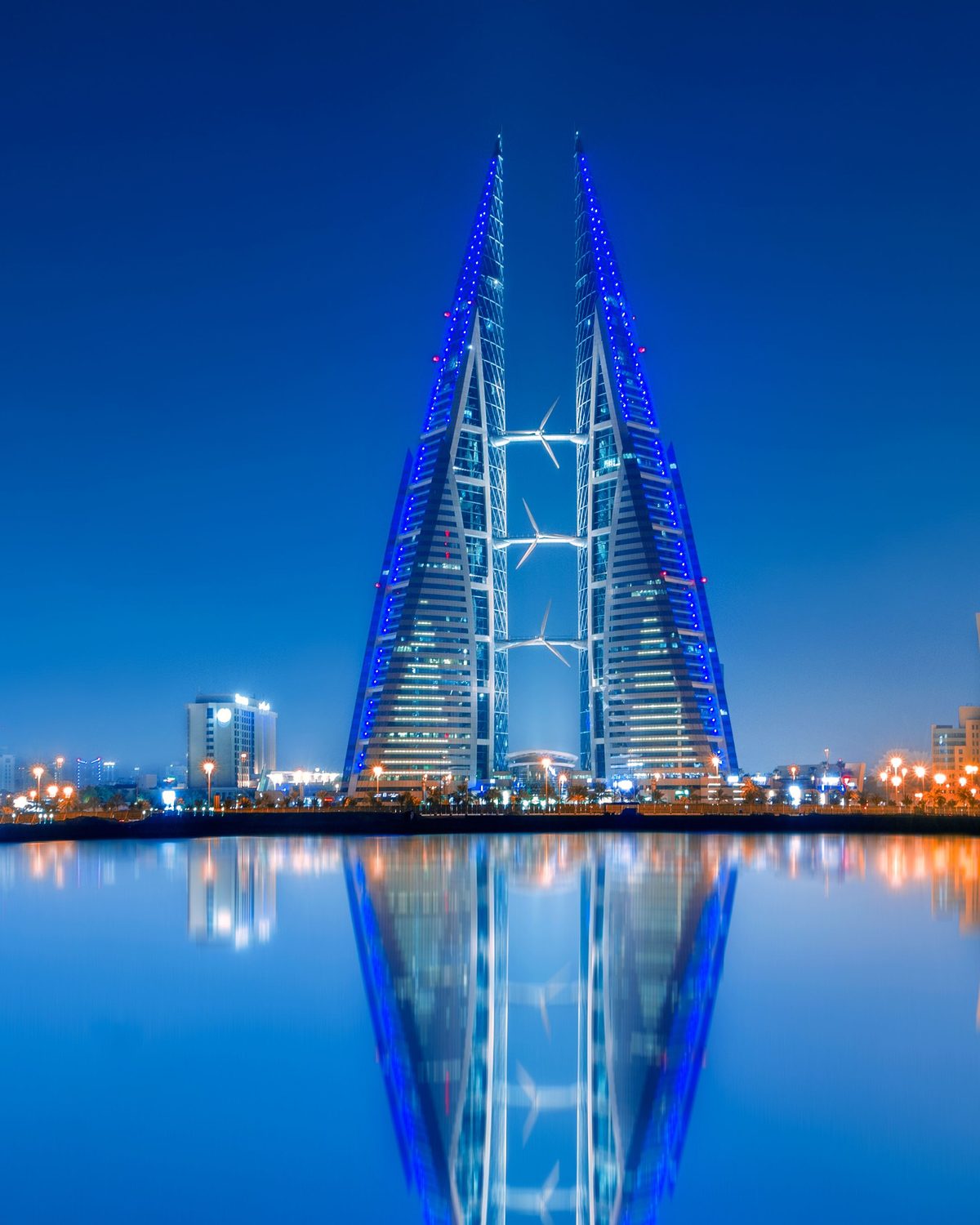 Bahrainin World Trade Centerin kaksoispurjeen muotoiset tornit sinisenä valaistuina iltahämärässä, heijastuen sataman tyyneen veteen.