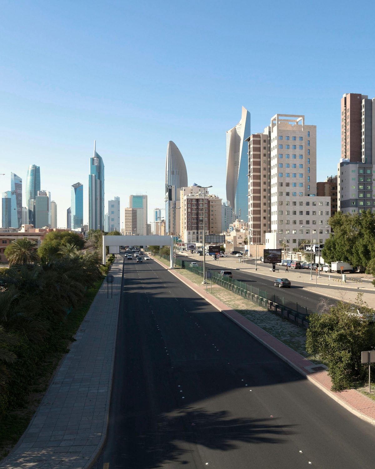 Kuwait Cityn moderni siluetti nähtynä palmuja reunustamalta puistokadulta kirkkaana päivänä.